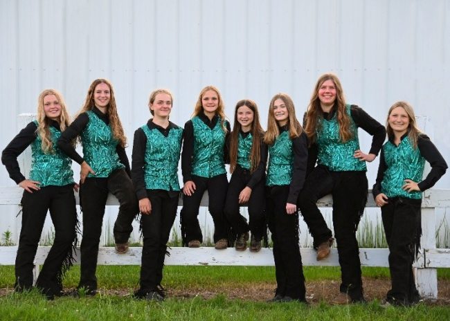 Team Glitter, a line of girls in green vests standing and sitting on a fence.